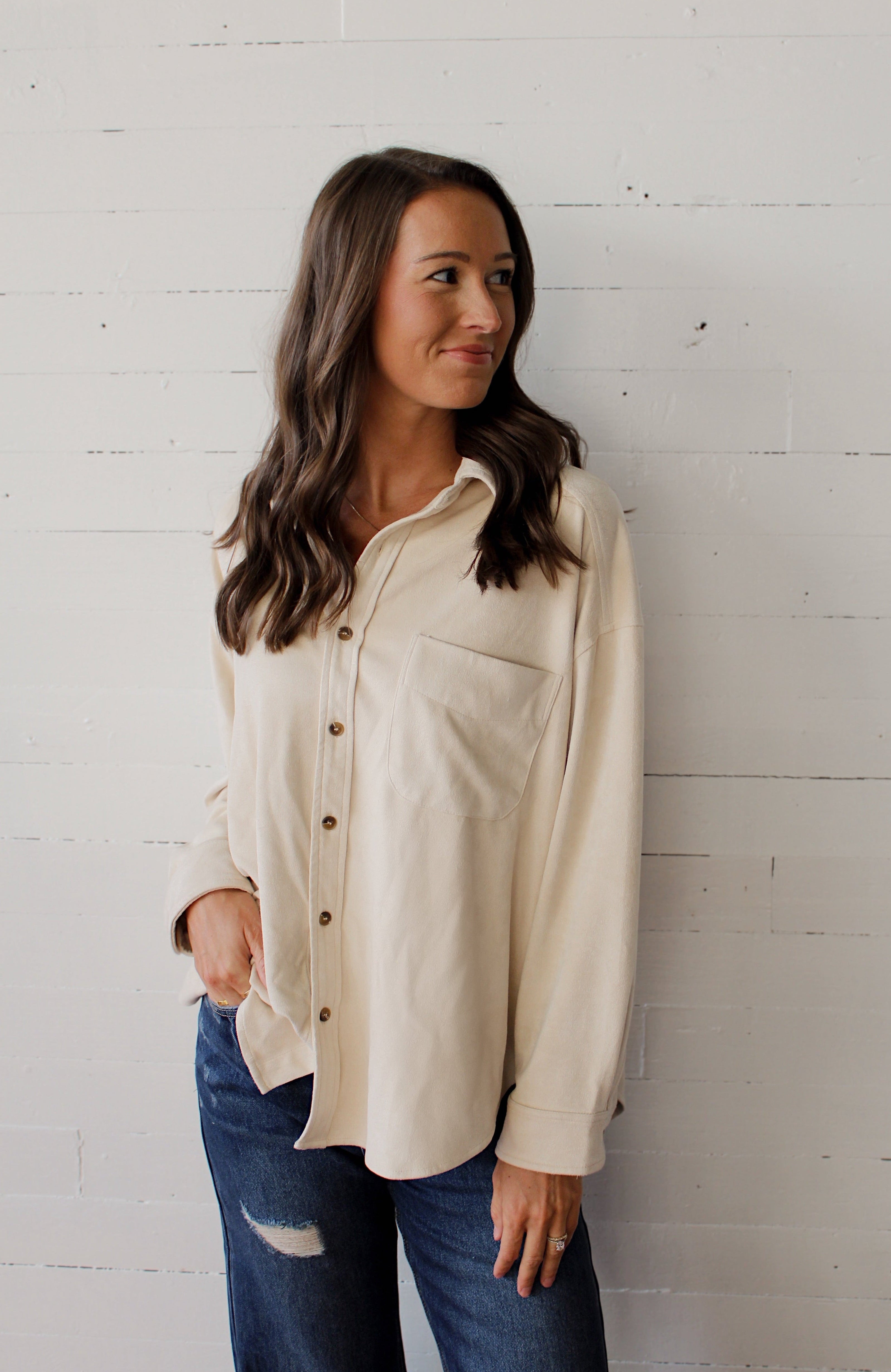 Woman wearing a cream button-up shirt and blue jeans against a white brick wall.