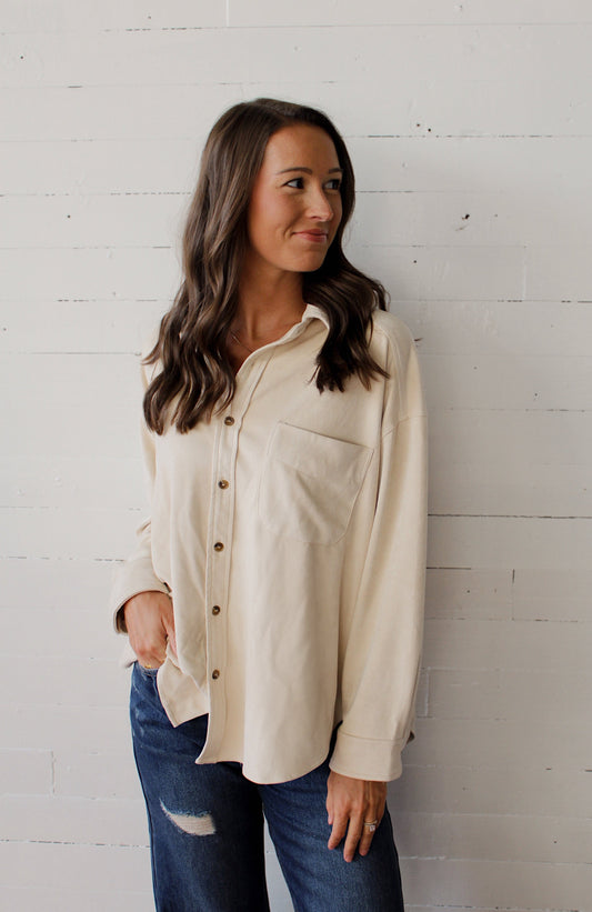 Woman wearing a cream button-up shirt and blue jeans against a white brick wall.
